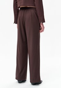 Persoon die donkerbruine wijde broek met elastische tailleband en een cropped bruin jasje met krijtstrepen draagt, staand tegen een witte achtergrond.