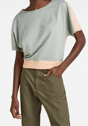 Person iført en lysegrå cropped skjorte med beige ærmer og kant, kombineret med højtaljede olivengrønne bukser med den ene hånd i lommen.