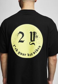 Juodas medvilnės marškinėliai su dideliu geltonu apvaliu grafiku ant nugaros, kuriame užrašyta "2Y" ir "f*ck your bad vibes" juodu fontu.