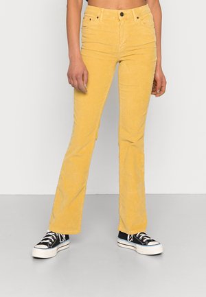 Calças de veludo amarelo com corte boca de sino, bolsos frontais, fecho de botão e uma textura ribbed distinta, combinadas com sapatilhas pretas de cano alto.
