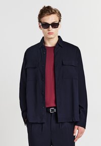 Camicia blu con bottoni e due tasche sul petto sopra una camicia borgogna a trama; abbinata a pantaloni scuri e occhiali da sole neri.