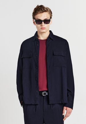 Chemise bleue marine à boutons avec deux poches sur la poitrine, portée par-dessus une chemise bordeaux texturée ; associée à un pantalon sombre et des lunettes de soleil noires.