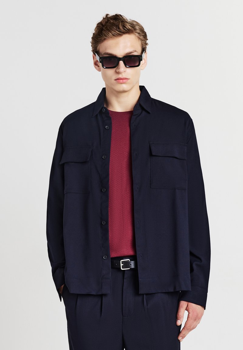 Camicia blu con bottoni e due tasche sul petto sopra una camicia borgogna a trama; abbinata a pantaloni scuri e occhiali da sole neri.