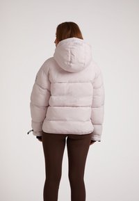 Lichtroze pufferjack met een capuchon, voorzien van horizontale gestikte delen en elastische manchetten. Subtiele branding op de achterkant.