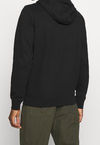 Sweat à capuche noir avec une texture douce, poignets et ourlet côtelés. Détails de coutures visibles le long des coutures. Porté avec un pantalon vert olive.