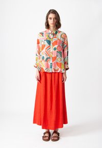 Kleurvol overhemd met geometrische patronen in roze, groen en oranje, gecombineerd met een rode maxi rok en bruine sandalen.
