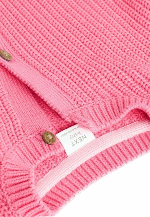 Rosa stickad babytröja med texturerad ribbning, som har två runda knappar och en synlig etikett på den inre fållen. Mjukt och mysigt tyg.