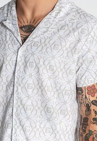 Kortom, witte blouse met korte mouwen, voorzien van een lichtgrijs bloempatroon, met knoopsluiting aan de voorkant en kraagdetails. Gestructureerde stof zichtbaar.
