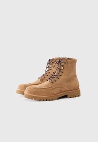 Botine din suede bej, cu șireturi maro, vârf rotund și talpă din cauciuc rezistentă. Prezintă ochiuri metalice și detalii subtile de cusătură.