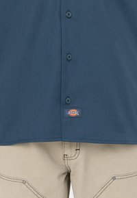 Camicia a maniche corte di colore blu scuro, realizzata in tessuto dalla texture liscia, con una fila di bottoni e un'etichetta con il logo Dickies nella parte inferiore.