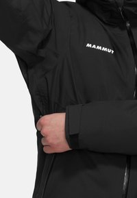 Giacca nera impermeabile con zip frontale, polsini regolabili e logo sul petto. La texture è liscia con un design leggero.