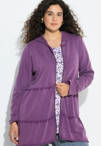 Žena s kudrnatými vlasy, která nosí zlaté kruhové náušnice a fialovou mikinu na zip s kapucí přes top s květinovým vzorem.