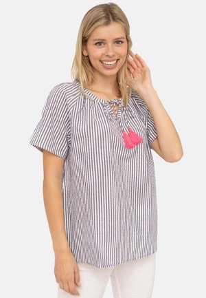 Lächelnde Frau mit blonden Haaren, die eine blau-weiß gestreifte Bluse mit rosa Quasten und weiße Hosen trägt und sich die Haare berührt.