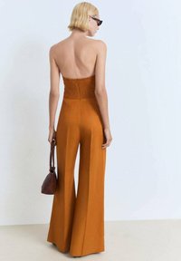 Strapless jumpsuit in een warme oranje tint met een smocked lijfje, brede pijpen en een soepele stof. Model houdt een kleine bruine tas vast.
