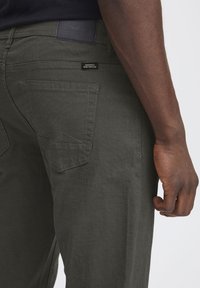 Pantalones verde oscuro hechos de un tejido duradero, con dos bolsillos traseros que tienen un detalle de solapa triangular y un acabado texturizado.