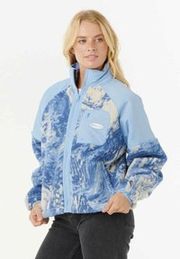 Blonde vrouw in een lichtblauwe en witte fleece jas met patroon, hoge kraag en ritszak op de borst, poseert met haar handen bij haar taille.