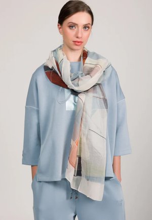 Mujer joven con blusa y pantalones sueltos en azul claro, llevando una bufanda grande con un patrón abstracto colorido alrededor del cuello, sobre un fondo liso.