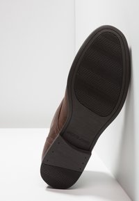 Zapato de cuero marrón con una suela de goma marrón texturizada. La suela presenta surcos horizontales para tracción y una marca en el talón.