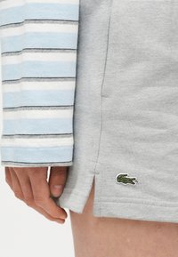 Χέρι δίπλα σε ανοιχτό γκρι σορτς με μικρό πράσινο λογότυπο κροκόδειλου· το μανίκι ενός μπλε, άσπρου και γκρι ριγέ πουκαμίσου καλύπτει μερικώς το χέρι.