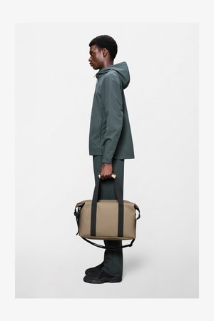 Beež tollekott mustade rihmade ja kangakahjuga, külje pealekandega. Modell kannab rohelist veekindlat jope ning sobivaid pükse ja musti jalatseid.