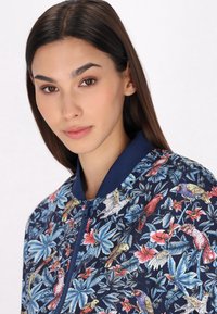 Giubbotto bomber blu navy con stampa floreale e di uccelli in colori vivaci, dotato di colletto a coste e chiusura con zip per un facile indosso.