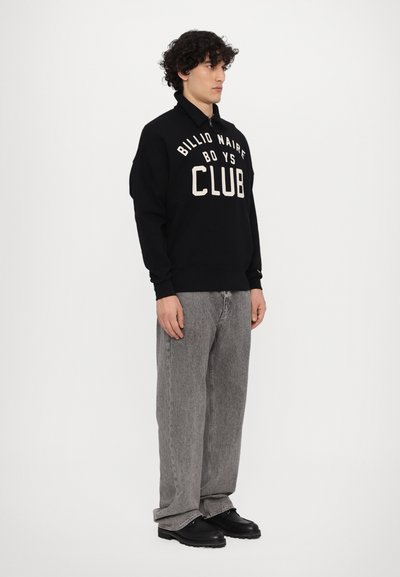 Μαύρη μπλούζα με κουκούλα "Billionaire Boys Club" σε κρεμ γράμματα, συνδυασμένη με γκρίζα φαρδιά τζιν παντελόνια και μαύρα παπούτσια, με σχέδιο κολάρου.