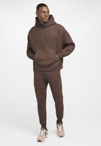 Brun hættejakke med frontlomme, afslappet pasform. Sammen med matchende tapered bukser med minimale sømme og branddetalje. Sko med lyse accenter.