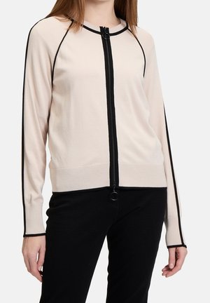 Žena nosící béžový cardigán na zip s černým lemováním podél zipu, rukávů a manžet, v kombinaci s černými kalhotami.