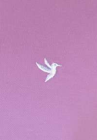Lila Stoff mit einer strukturierten Oberfläche zeigt ein weiß besticktes Kolibri-Logo in der unteren rechten Ecke.