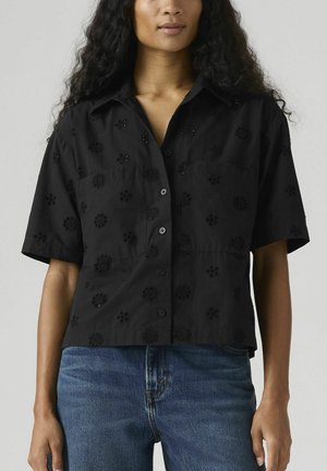 Frau trägt ein schwarzes kurzärmeliges Hemd mit Knöpfen und floralen Locher-Mustern sowie blaue Jeans vor einem einfarbigen Hintergrund.