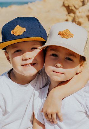 Zwei Kinder mit Mützen: eine marineblaue mit einem gelben Schirm und Logo, die andere beige mit einem Netzrücken und einem ähnlichen Logo. Beide tragen weiße Hemden.