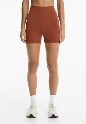 Shorts a vita alta di colore ruggine con una texture liscia e un design a cucitura geometrica, abbinati a scarpe da ginnastica chiare e calzini bianchi.