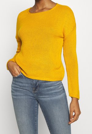 Frau trägt einen leuchtend gelben Strickpullover mit langen Ärmeln und hellblauen, enganliegenden Jeans, posiert mit einer Hand in der Tasche.