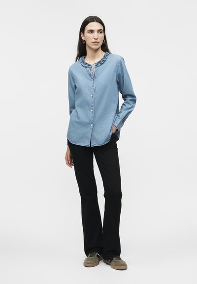 Γυναίκα που στέκεται με το αριστερό χέρι στην τσέπη, φορώντας μπλε μπλούζα με βολάν στο γιακά, μαύρο παντελόνι και ελιές πράσινα αθλητικά παπούτσια σε λευκό φόντο.