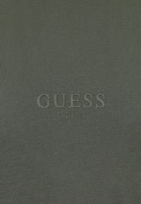 Ciemnozielony materiał z tłoczonym w dopasowanym kolorze napisem "GUESS EST 1981" na środku.