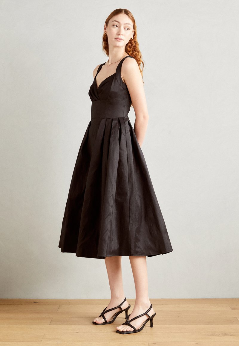 Black knee-length dress with a fitted bodice, pleated skirt, wide straps, and a smooth texture. Paired with black strappy heels.