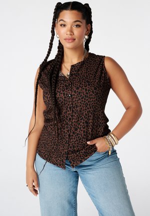 Femme aux longs cheveux tressés portant une chemise sans manches marron et noire à imprimé léopard avec boutons et un jean bleu clair avec des bracelets au poignet droit.
