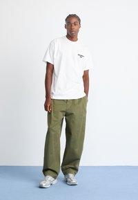 Witte katoenen t-shirt met zwart logo, gecombineerd met olijfgroene wide-leg broek en lichtgrijze sneakers. Glad textuur, relaxte pasvorm.