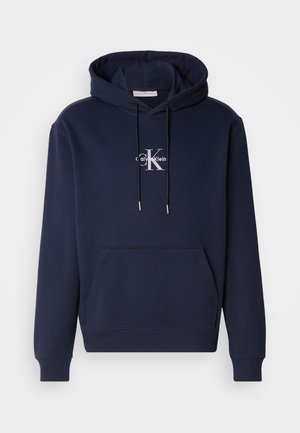 Mornarsko plavi dukserica sa prednjim džepom, kapuljačom na vezanje i bijelim logom Calvin Klein. Izrađena od mekog materijala s rebrastim manžetama i donjim rubom.