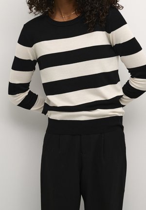 Osoba stojící s rukama v bok, oblečená v černobílém pruhovaném svetru s dlouhým rukávem a černých kalhotách.