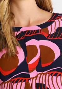 Blouse en coton colorée avec un col rond, des motifs abstraits audacieux en rose et marron, et un tissu texturé avec un léger brillant.