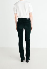 Pantalon évasé en velours côtelé vert à finition texturée, avec deux poches arrière et une petite étiquette logo près de la ceinture.