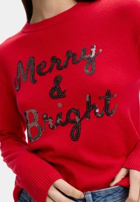 Maglione rosso lavorato a maglia con polsini a coste, caratterizzato dalla scritta "Merry & Bright" in paillettes nere. La vista ravvicinata mette in evidenza la texture e i dettagli del design.