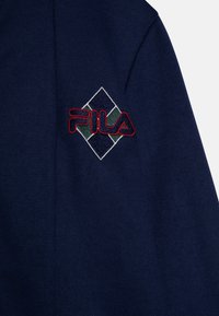 Il tessuto della giacca navy presenta un logo ricamato "FILA" rosso all'interno di un design a diamante, arricchito da dettagli verdi e blu.
