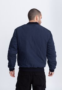 Marineblauwe bomberjack met een ronde kraag, geribbelde manchetten en een geribde tailleband. Kenmerkt zich door een gestructureerde stof en een strakke, aansluitende silhouet.