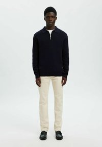 Marineblauer Strickpullover mit Reißverschluss am Kragen, kombiniert mit hellbeigen, gerade geschnittenen Hosen und schwarzen Lederschuhen. Schlichter, strukturierter Look.