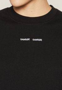 Černé bavlněné tričko s kulatým výstřihem, na hrudi dvakrát s bílým vyšívaným textem "SAMSDØ". Hladká textura, standardní střih.