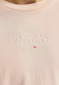 Šviesiai rožinė medvilnės marškinėliai su apvalia iškirpte, ant kurių yra įspaustas baltas tekstas "JACK & JONES PREMIUM" ir mažas prekių ženklų simbolis.