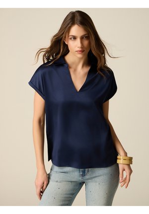 Donna con capelli castani sciolti indossa una blusa blu navy a maniche corte con scollo a V e jeans azzurri con piccoli dettagli decorativi.