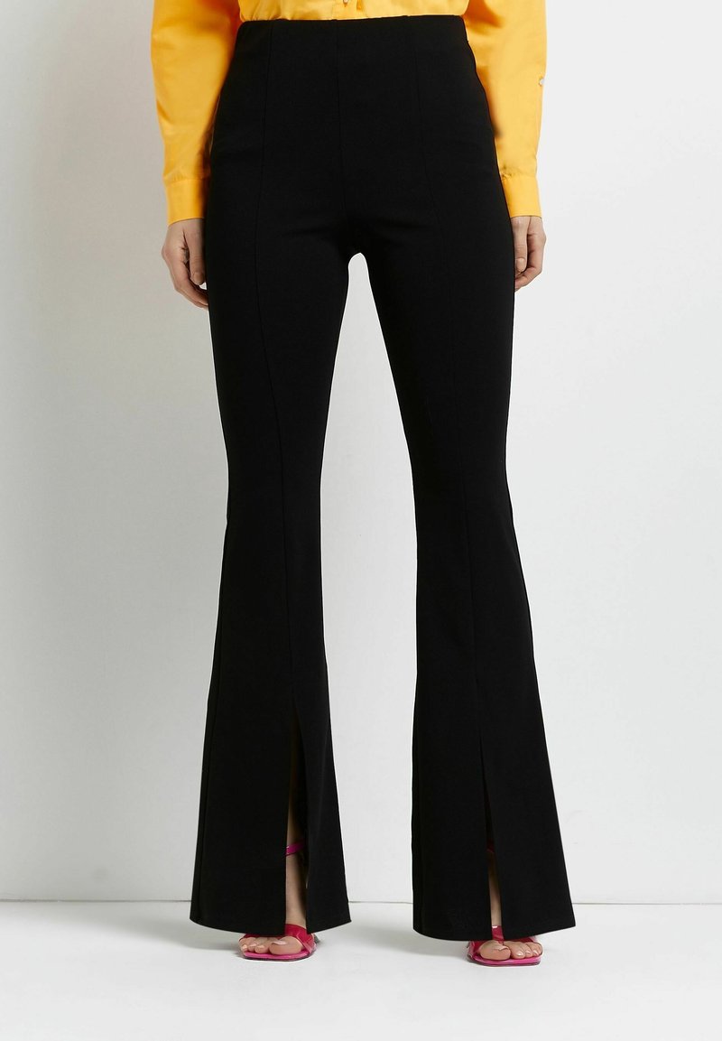 Mujer con pantalones negros acampanados de talle alto con aberturas frontales, combinados con una blusa amarilla de manga larga y sandalias rosas abiertas en los dedos.